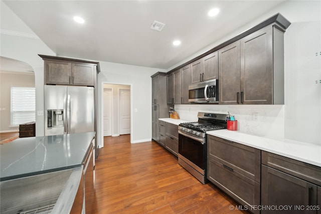 kitchen featuring dark brown cabinetry, stainless steel appliances, dark hardwood / wood-style floors, backsplash, and ornamental molding