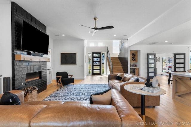 living area featuring crown molding, light wood finished floors, stairway, a brick fireplace, and ceiling fan