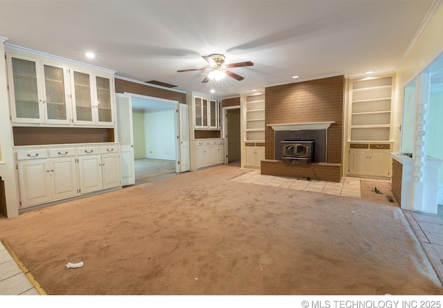 unfurnished living room with a wood stove, built in shelves, ceiling fan, ornamental molding, and light colored carpet