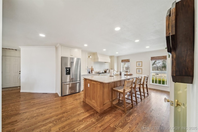 kitchen with high end fridge, custom range hood, a center island with sink, white cabinets, and hardwood / wood-style floors