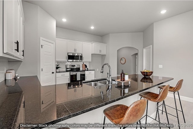 kitchen with arched walkways, a sink, white cabinetry, appliances with stainless steel finishes, and dark stone counters