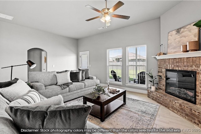 living room with arched walkways, visible vents, light wood-style flooring, a ceiling fan, and a brick fireplace