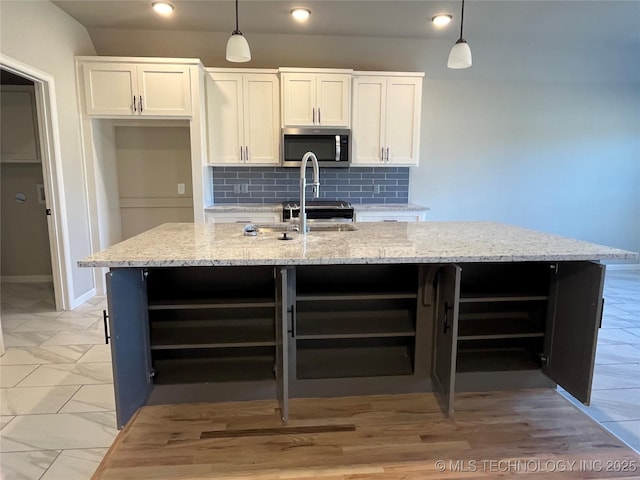 kitchen featuring light stone counters, a kitchen island with sink, white cabinetry, backsplash, and stainless steel microwave