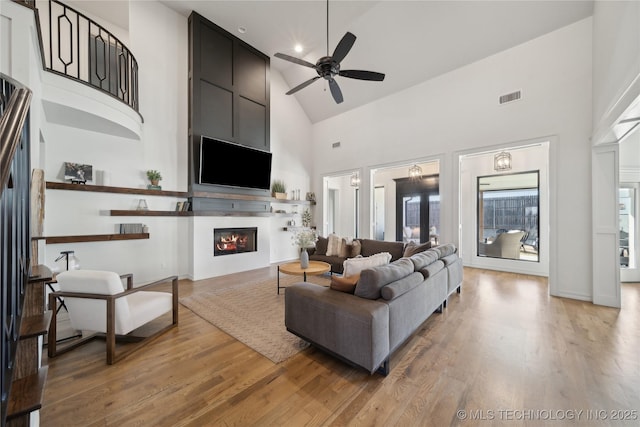 living room with light wood-type flooring, ceiling fan, a large fireplace, and high vaulted ceiling