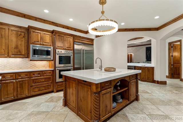 kitchen featuring hanging light fixtures, built in appliances, an island with sink, decorative backsplash, and ornamental molding