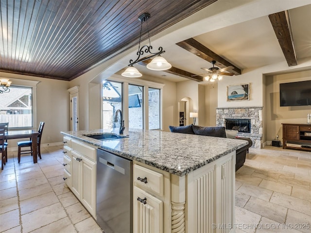 kitchen with sink, a stone fireplace, stainless steel dishwasher, an island with sink, and pendant lighting