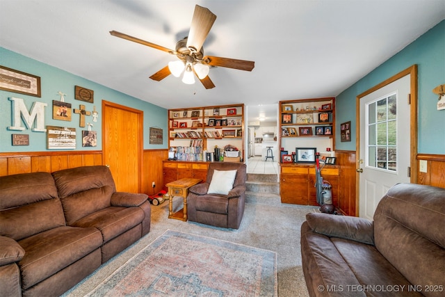 living room featuring carpet flooring, built in shelves, wooden walls, and ceiling fan