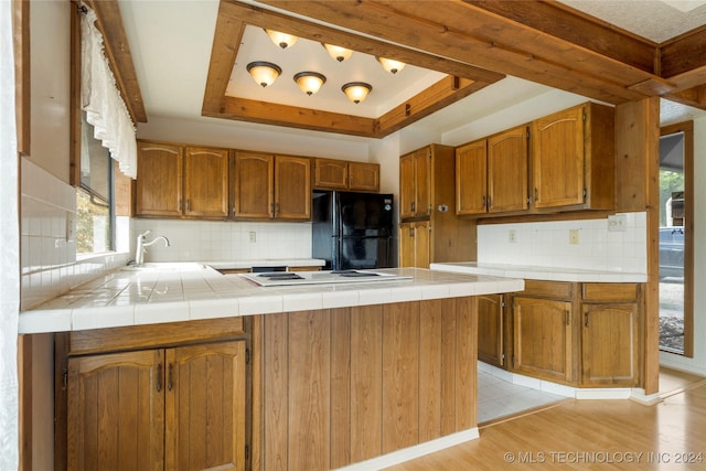 kitchen with tile counters, tasteful backsplash, black fridge, a tray ceiling, and light wood-type flooring