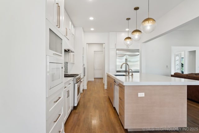 kitchen with wood-type flooring, built in appliances, decorative light fixtures, a kitchen island with sink, and white cabinets