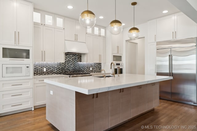 kitchen with decorative light fixtures, white cabinets, a kitchen island with sink, built in appliances, and custom range hood