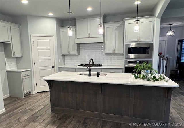 kitchen with light stone counters, stainless steel appliances, dark wood-type flooring, hanging light fixtures, and an island with sink