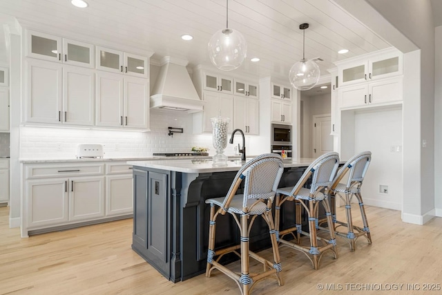 kitchen featuring custom exhaust hood, decorative light fixtures, white cabinets, light hardwood / wood-style floors, and an island with sink