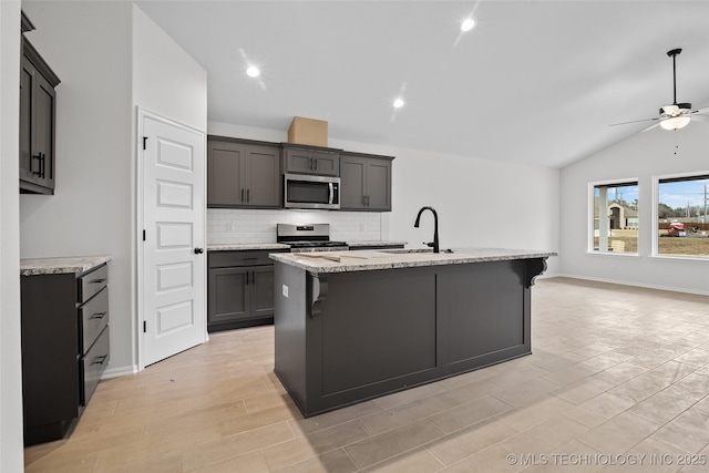 kitchen featuring sink, light stone counters, an island with sink, vaulted ceiling, and appliances with stainless steel finishes