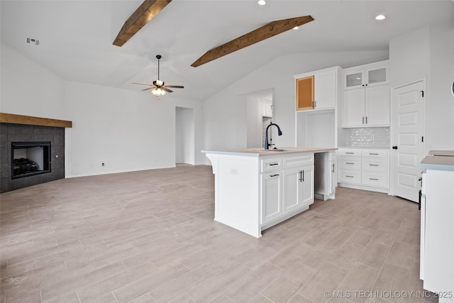 kitchen featuring tasteful backsplash, an island with sink, ceiling fan, a tiled fireplace, and white cabinets