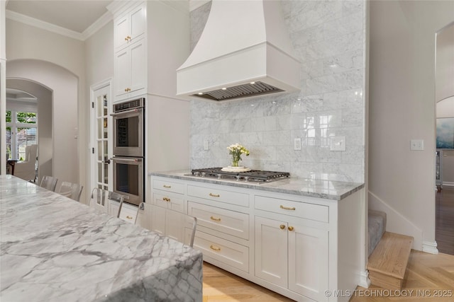 kitchen with light stone counters, premium range hood, backsplash, white cabinets, and appliances with stainless steel finishes