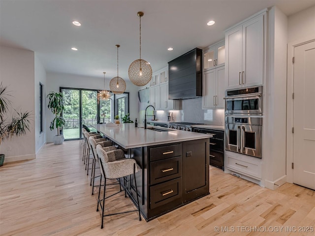 kitchen with premium range hood, double oven, sink, a center island with sink, and hanging light fixtures