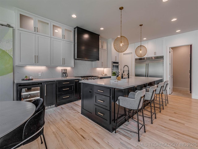 kitchen featuring appliances with stainless steel finishes, custom exhaust hood, a center island with sink, decorative light fixtures, and white cabinetry