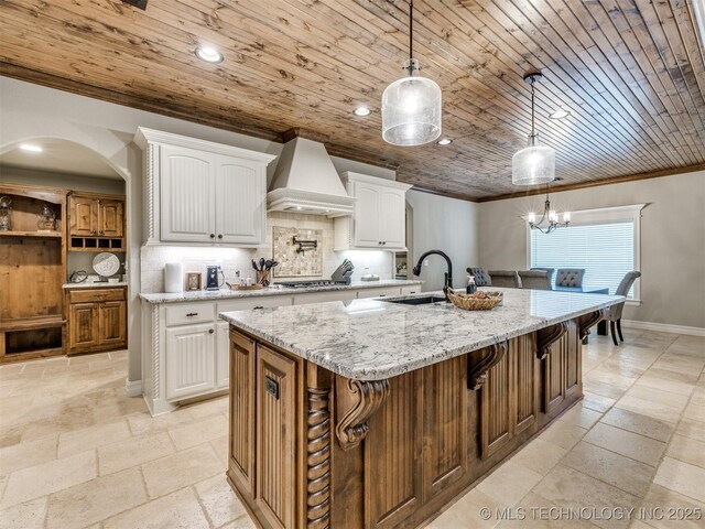 kitchen featuring white cabinetry, custom range hood, decorative light fixtures, and an island with sink