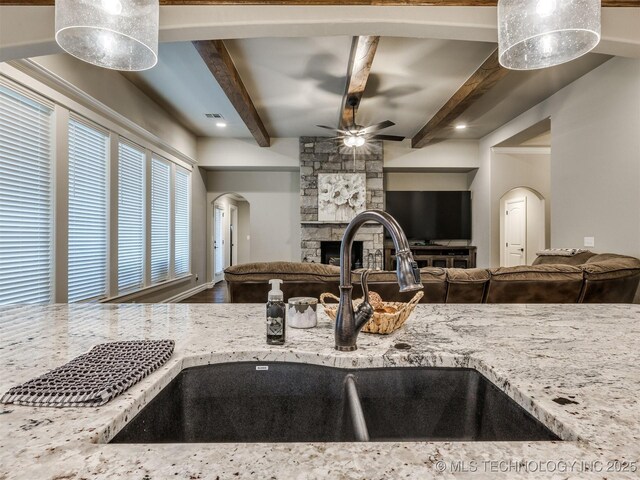 kitchen with a stone fireplace, beamed ceiling, sink, ceiling fan, and light stone countertops