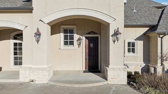 property entrance with stucco siding, roof with shingles, and stone siding