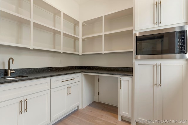 kitchen featuring built in microwave, sink, dark stone counters, white cabinets, and light wood-type flooring