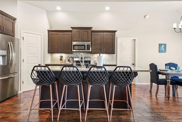 kitchen with appliances with stainless steel finishes, light stone counters, dark brown cabinets, and an island with sink