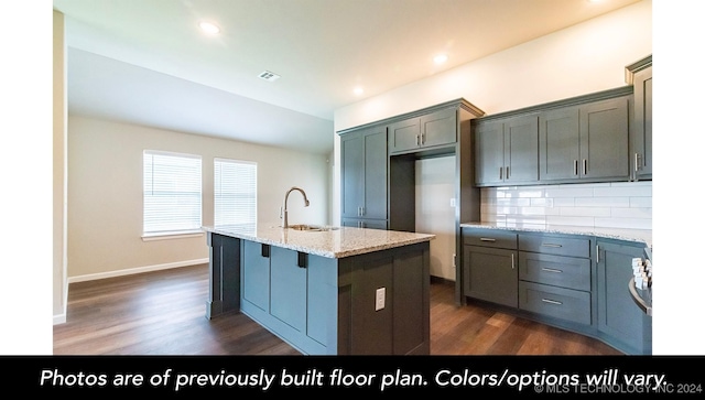 kitchen with light stone countertops, tasteful backsplash, dark wood-type flooring, sink, and a center island with sink