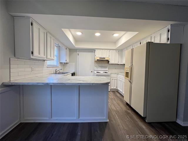 kitchen with white appliances, dark wood-type flooring, white cabinets, a tray ceiling, and kitchen peninsula