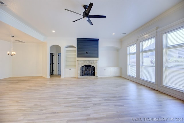 unfurnished living room with ceiling fan with notable chandelier, a stone fireplace, built in shelves, light wood-type flooring, and ornamental molding