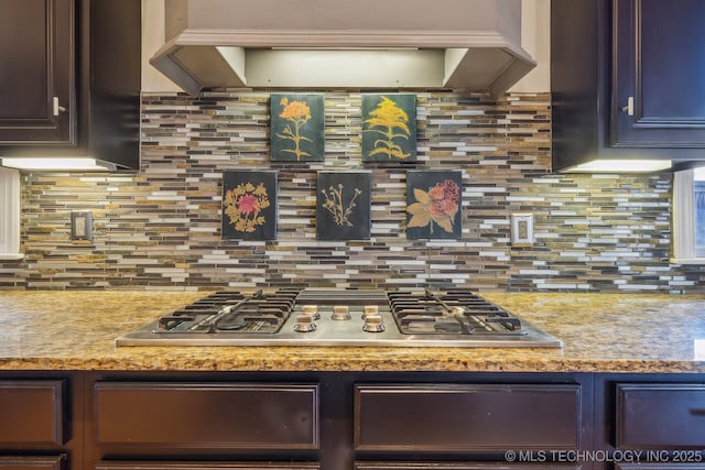 kitchen featuring stainless steel gas stovetop, premium range hood, decorative backsplash, light stone countertops, and dark brown cabinets