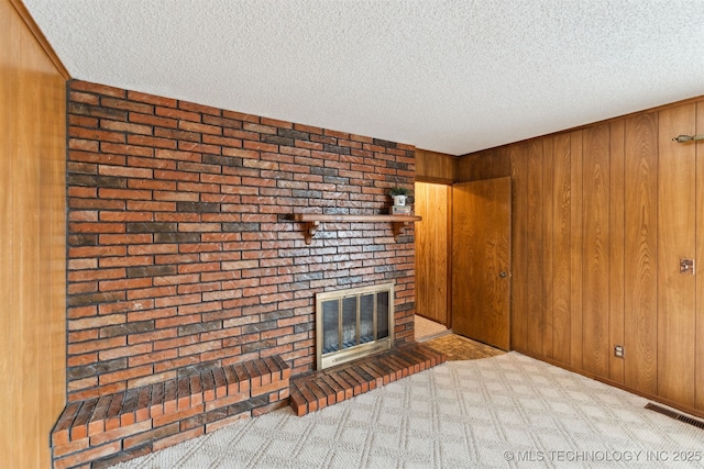 unfurnished living room with wooden walls, light colored carpet, a textured ceiling, and a brick fireplace