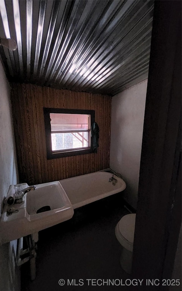 bathroom with a tub to relax in, toilet, wooden walls, and sink