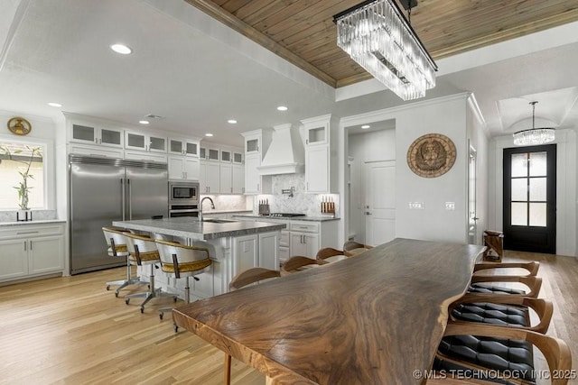 kitchen with custom exhaust hood, a breakfast bar, built in appliances, a center island with sink, and white cabinetry