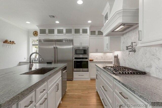 kitchen with custom range hood, built in appliances, white cabinetry, and sink