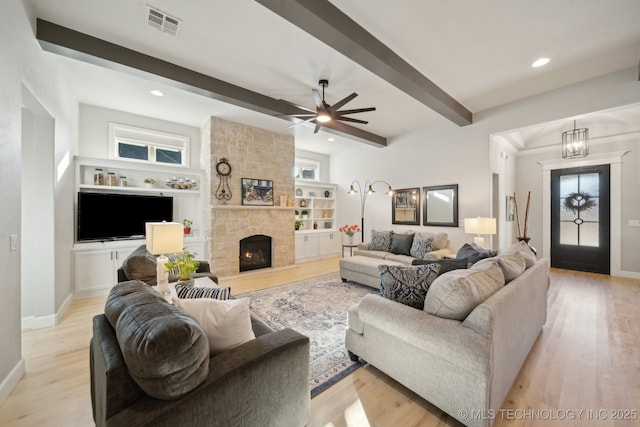 living room featuring beamed ceiling, ceiling fan with notable chandelier, a stone fireplace, and light wood-type flooring