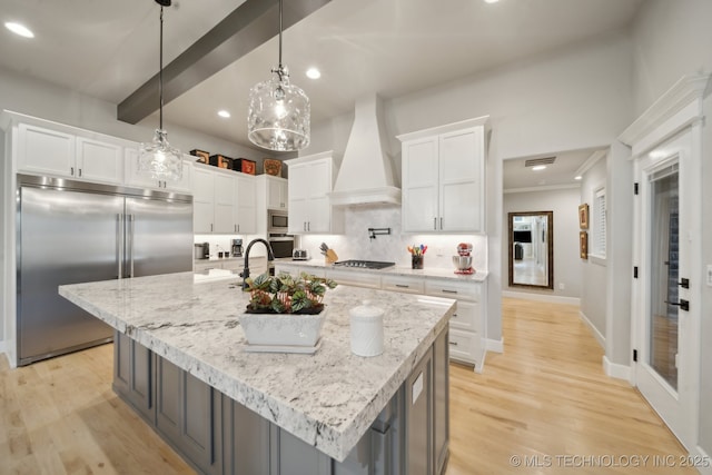 kitchen with custom exhaust hood, a kitchen island with sink, built in appliances, beamed ceiling, and white cabinets