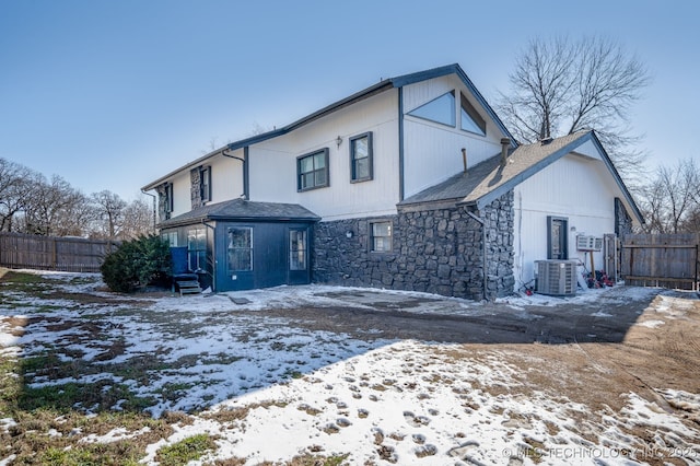 snow covered house featuring stone siding, central AC unit, and fence