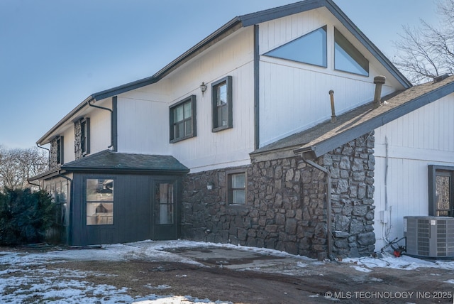 snow covered property with stone siding, roof with shingles, and central air condition unit