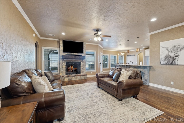 living room featuring ceiling fan with notable chandelier, a textured ceiling, crown molding, hardwood / wood-style flooring, and a stone fireplace