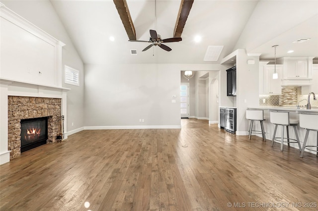 unfurnished living room with ceiling fan, hardwood / wood-style floors, beamed ceiling, a stone fireplace, and beverage cooler