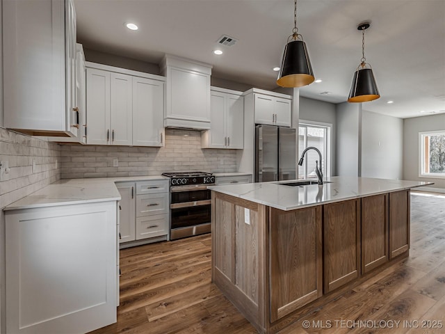 kitchen featuring range with two ovens, an island with sink, white cabinets, and stainless steel refrigerator