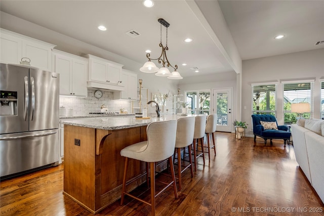 kitchen featuring light stone countertops, a center island with sink, decorative light fixtures, stainless steel fridge with ice dispenser, and white cabinetry