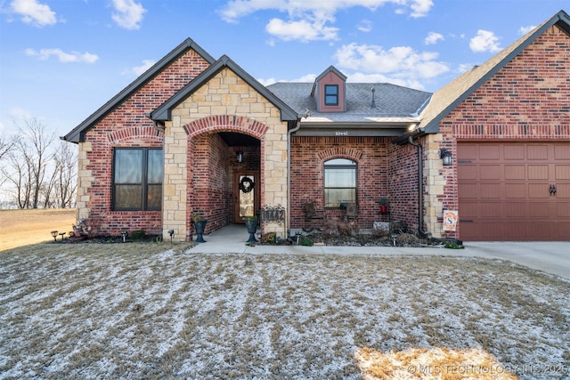 french country home featuring brick siding, driveway, an attached garage, and roof with shingles