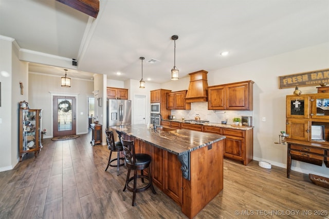 kitchen with pendant lighting, custom range hood, a kitchen island with sink, and appliances with stainless steel finishes