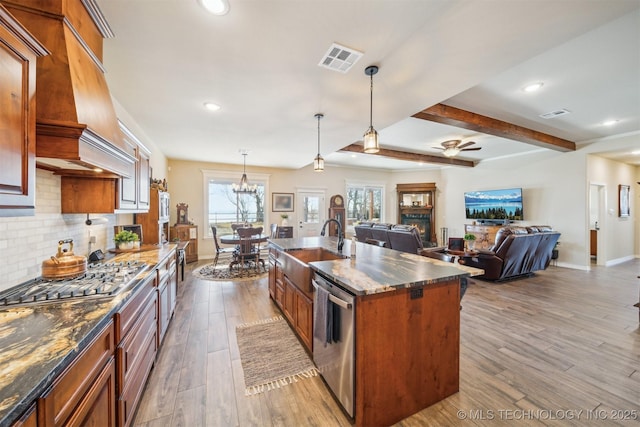 kitchen with appliances with stainless steel finishes, custom range hood, a kitchen island with sink, beamed ceiling, and hanging light fixtures