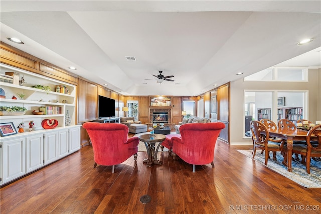 living area with built in shelves, dark wood-style floors, visible vents, a ceiling fan, and a glass covered fireplace