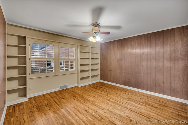 unfurnished room with ceiling fan, wood walls, light hardwood / wood-style floors, crown molding, and built in shelves