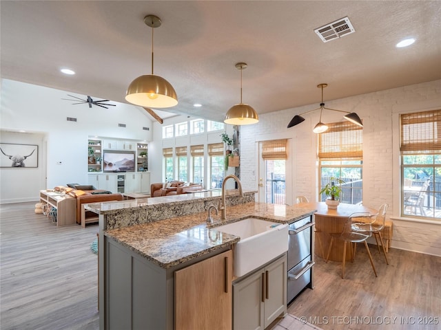 kitchen featuring light stone countertops, brick wall, a kitchen island with sink, sink, and pendant lighting