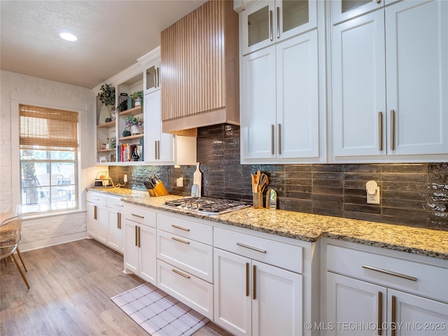 kitchen featuring light stone counters, backsplash, stainless steel gas stovetop, white cabinets, and light wood-type flooring