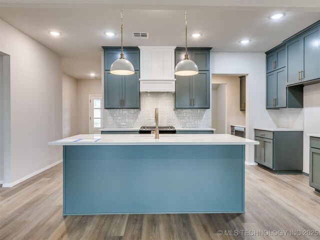 kitchen with sink, tasteful backsplash, decorative light fixtures, light wood-type flooring, and an island with sink